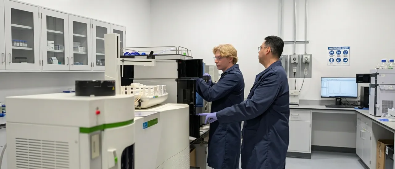 Two AMP Tech technicians in navy lab coats working on a PerkinElmer ICP-MS in the Longwood, Florida lab, a chromatogram on the monitor behind them
